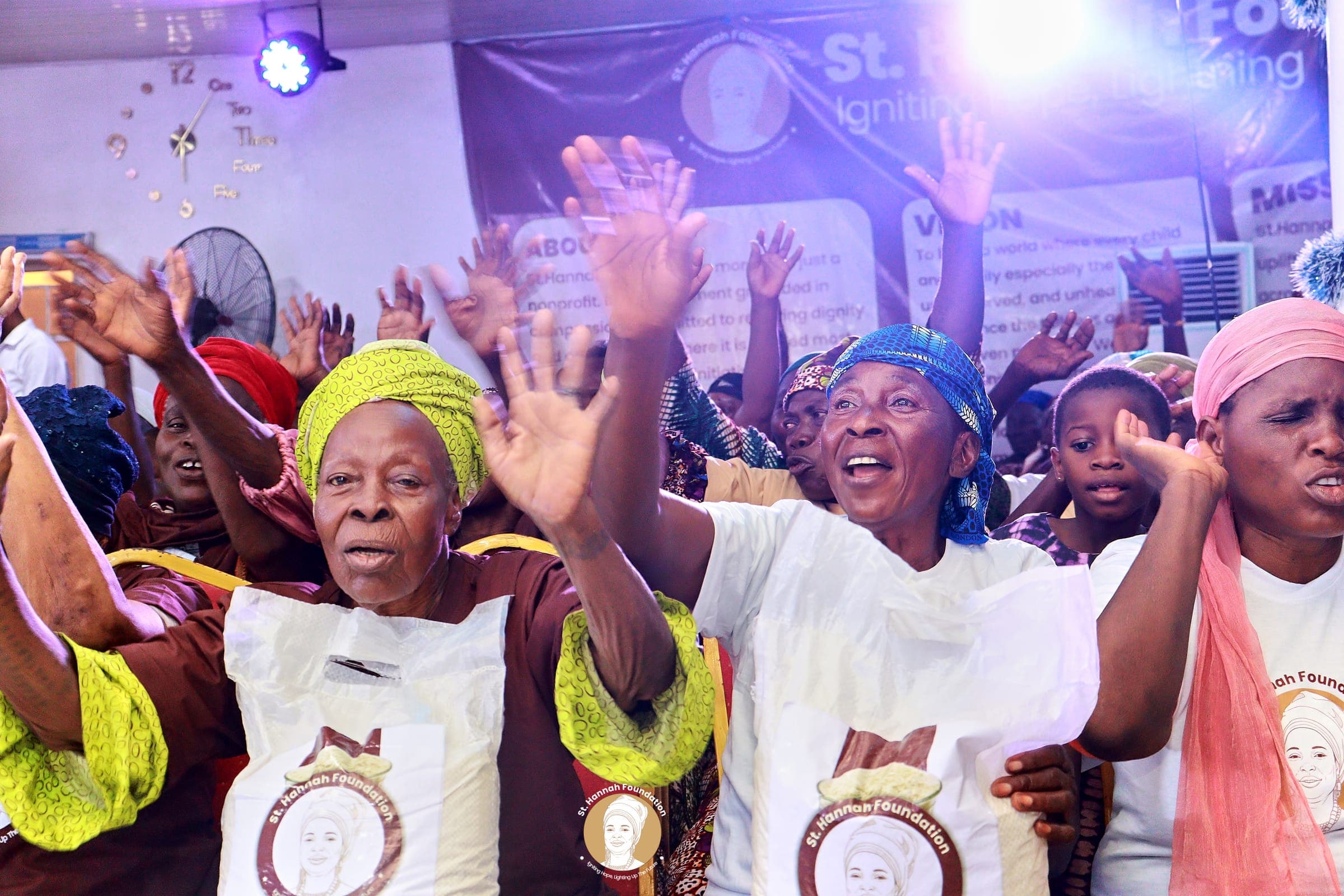 Older women in a hall raise their hands and sing, some wearing white bags printed with a portrait logo.