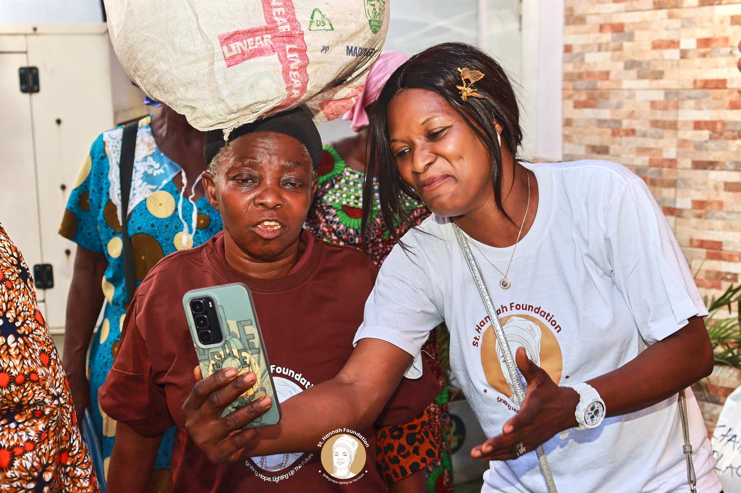 A volunteer takes a selfie with a smiling widow carrying a bag on her head.